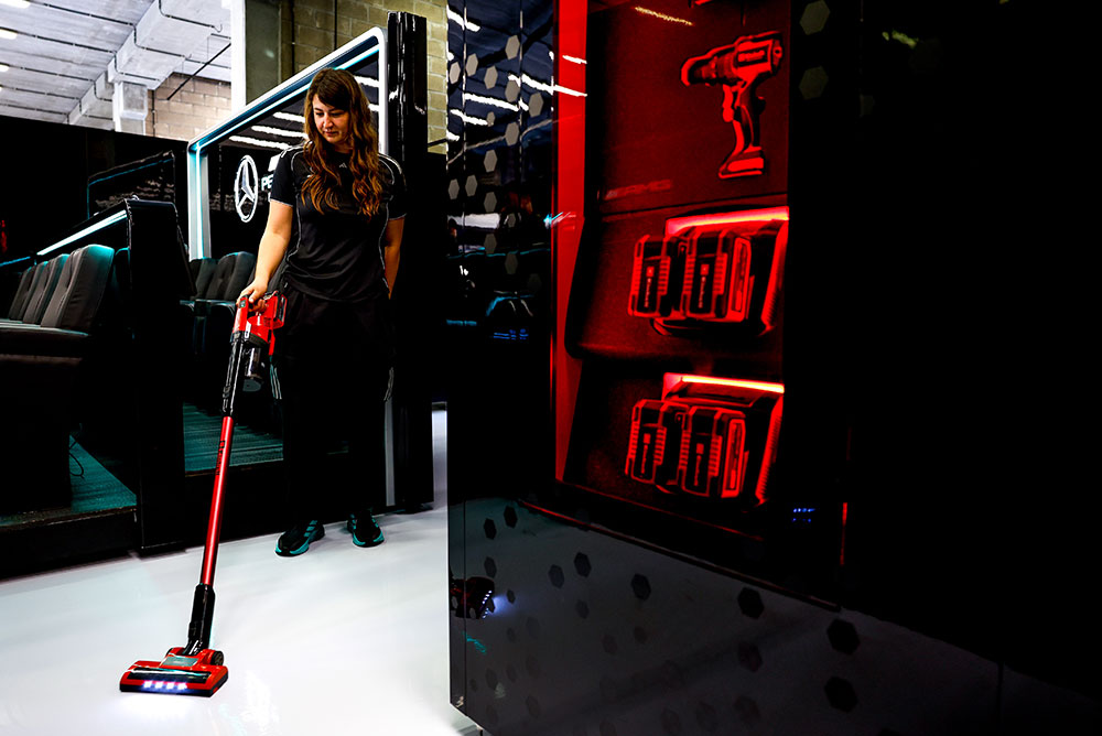 A woman from the Mercedes-AMG PETRONAS Formula One Team is cleaning a shiny white floor using an Einhell cordless vacuum cleaner. On the right, red-lit shelves showcase multiple Einhell Power X-Change batteries and a cordless drill.
