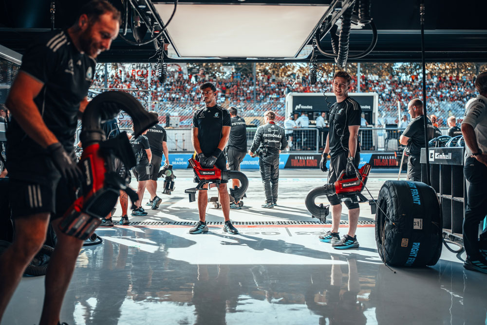 Mercedes-AMG PETRONAS Formula One Team mechanics are preparing in the garage with Einhell cordless blowers before a pit stop. In the background, the grandstands are filled with fans.