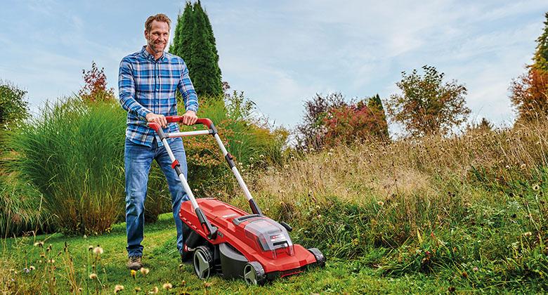 A man is mowing an uneven lawn with tall grass at the edge using a red cordless lawn mower.
