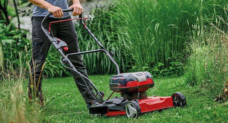A man is mowing tall grass with a red cordless lawn mower on a wild, natural lawn.
