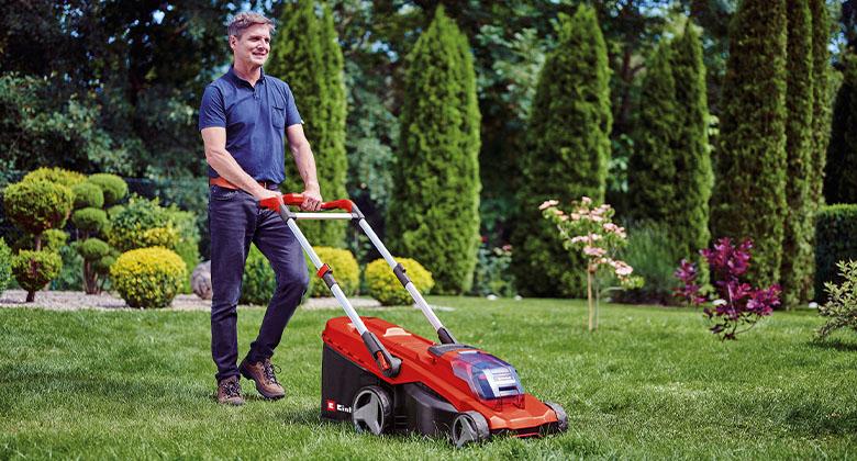 A man is mowing the lawn with a red cordless lawn mower in a well-maintained garden with ornamental shrubs and trees.