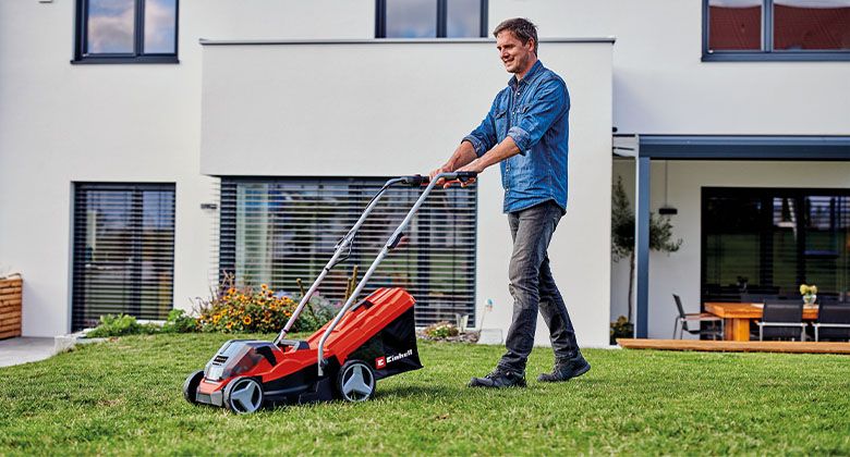 A man is mowing the lawn with a red cordless lawn mower in front of a modern white house.