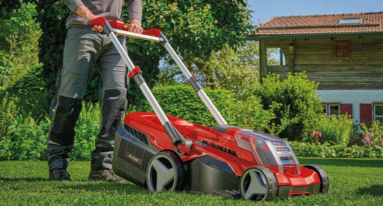 A man is mowing the lawn with a red cordless lawn mower in front of a traditional house with a garden.