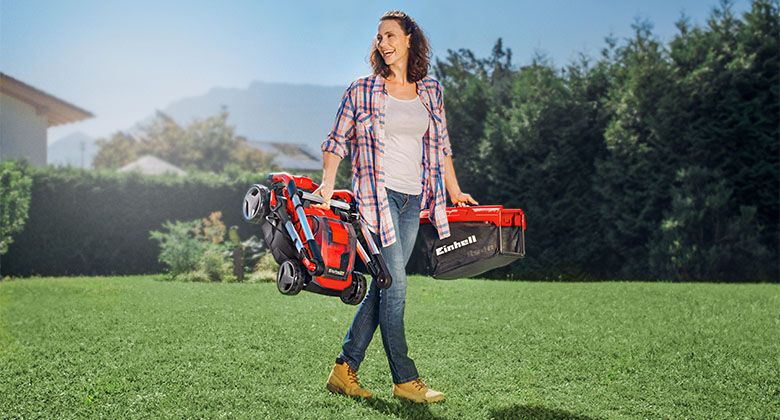 A woman is carrying a folded red cordless lawn mower in one hand and the grass collection bag in the other while standing on a well-kept lawn.